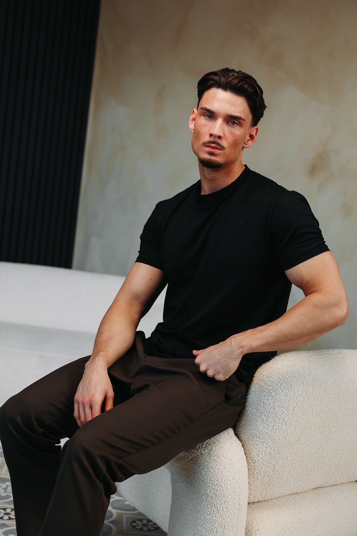 Model in het zwarte Shiny Essential Shirt, zittend op de leuning van een witte bank