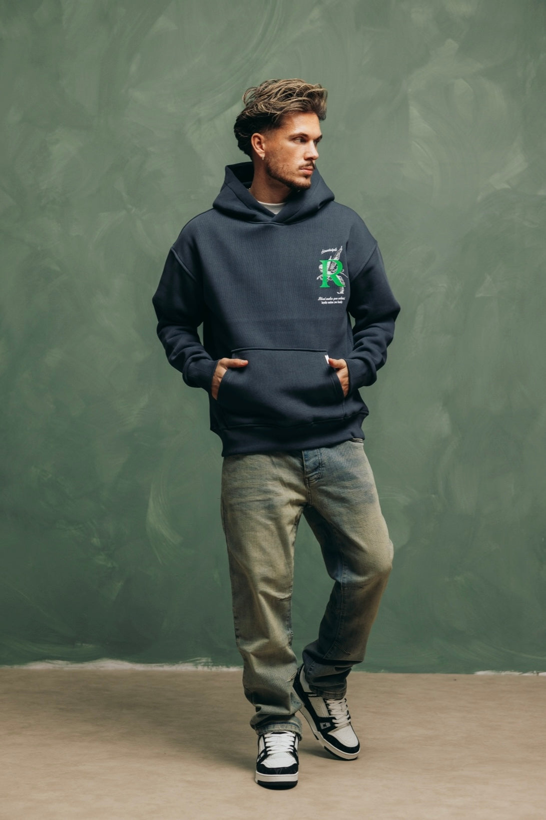 Model in de Artemis Hoodie met lichte jeans en sneakers, tegen een groene muur