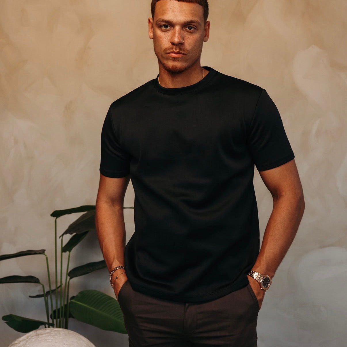 Model in het zwarte Gloss Essential Shirt met een donkere broek, tussen de planten